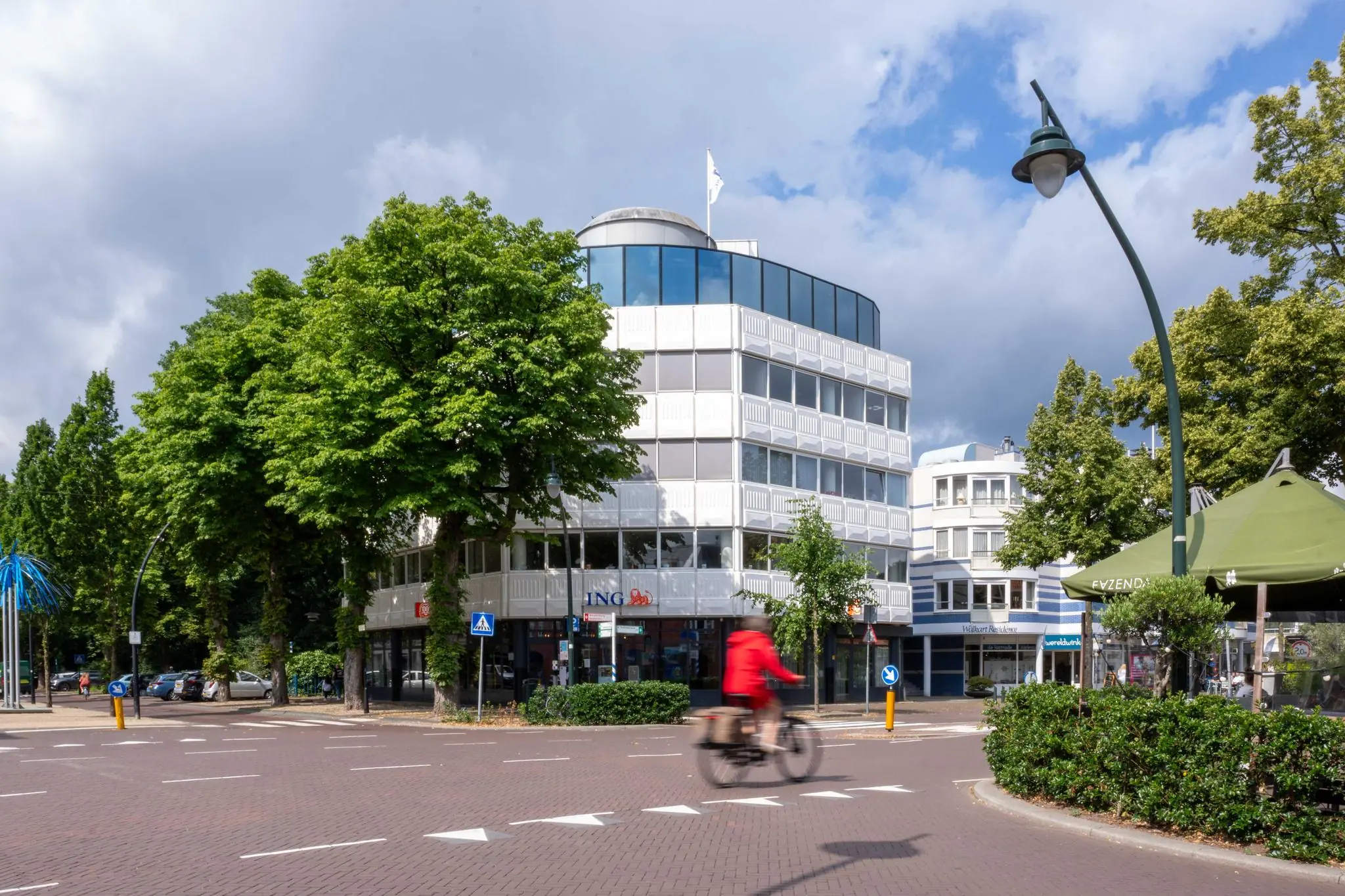 Fietser in rode jas rijdt langs het ING-gebouw aan de 1e Hogeweg in een groene, stedelijke omgeving.